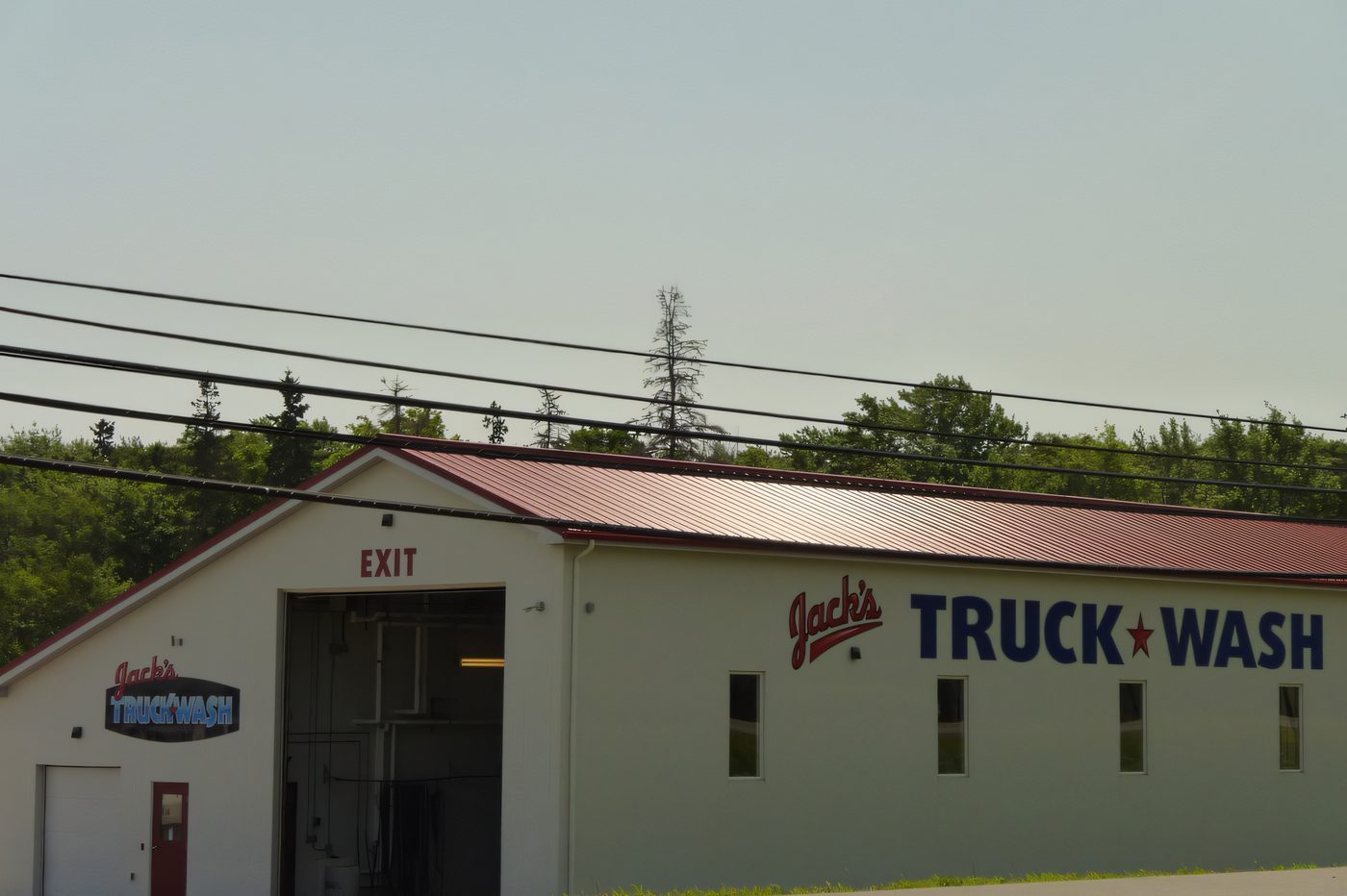 A white steel truck wash building with a red roof and a wide bay door, seen from the road