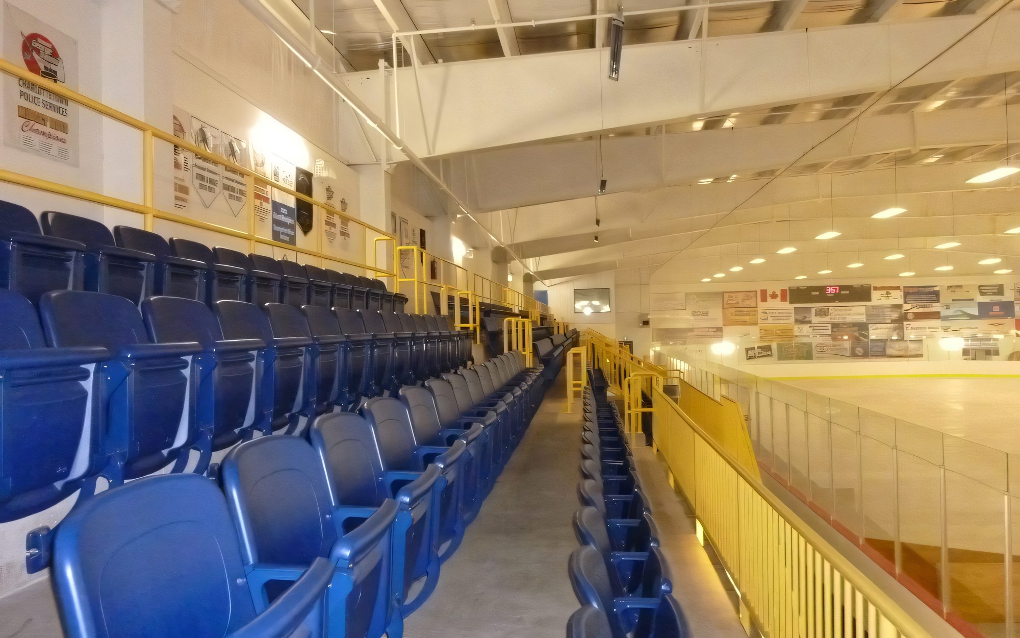 Inside a community arena: rows of blue seats behind yellow railings, the ice surface and the steel roof structure lit for the evening